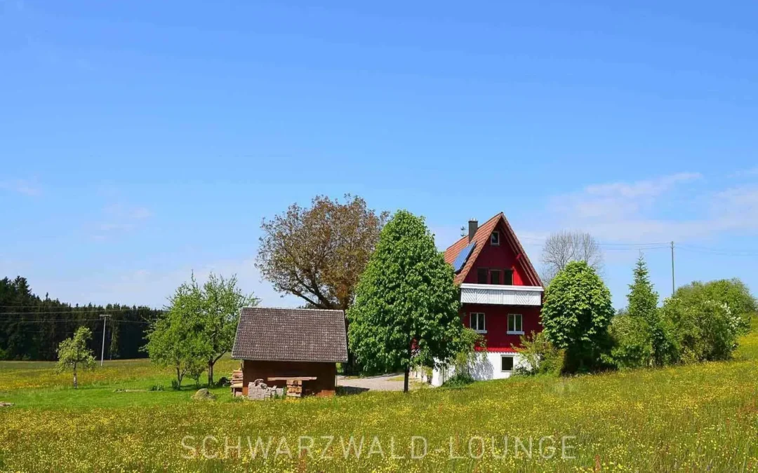 Rotes Ferienhaus Brestenberg inmitten weiter Wiesen mit Bäumen und klarblauem Himmel.