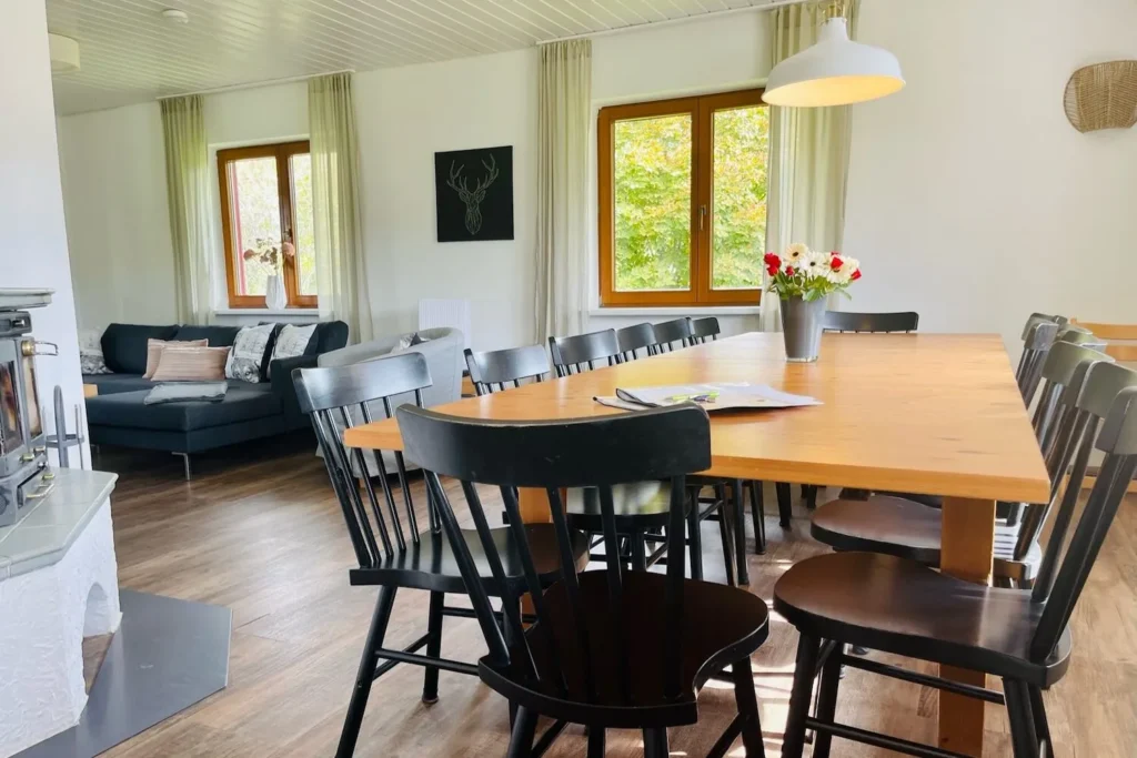 Helles Esszimmer mit großem Holztisch, schwarzen Stühlen und angrenzendem Wohnbereich mit Kaminofen.