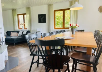 Helles Esszimmer mit großem Holztisch, schwarzen Stühlen und angrenzendem Wohnbereich mit Kaminofen.