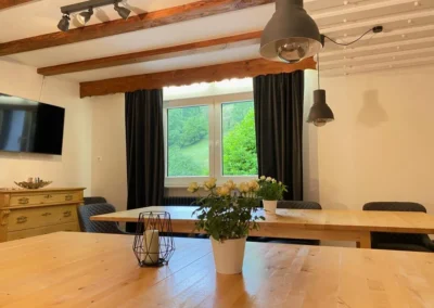 Aufenthaltsraum der Gruppenunterkunft im Schwarzwald mit großen Holztischen, Fensterblick ins Grüne und modernen Hängelampen.