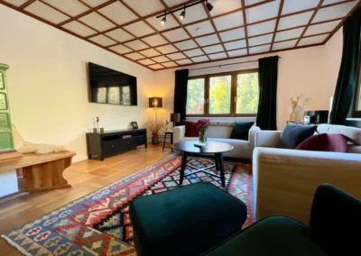 Gemütliches Wohnzimmer mit zwei Sofas, buntem Teppich, Kachelofen und großem Fenster in der Gruppenunterkunft Schwarzwald.