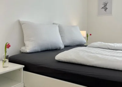 Helles Schlafzimmer der Ferienwohnung Westweg mit Doppelbett, frischen Blumen und modernem Nachttisch.