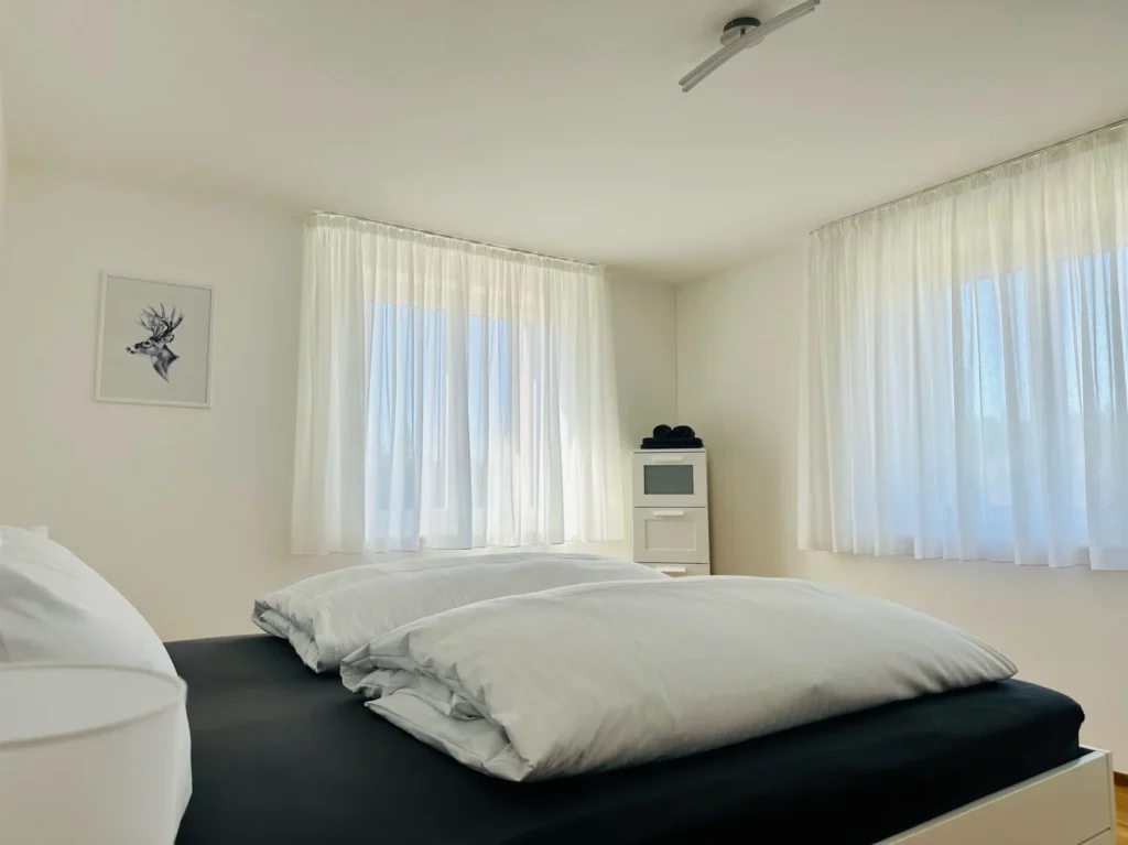 Schlafzimmer der Ferienwohnung Westweg mit großen Fenstern, hellen Vorhängen und Doppelbett.