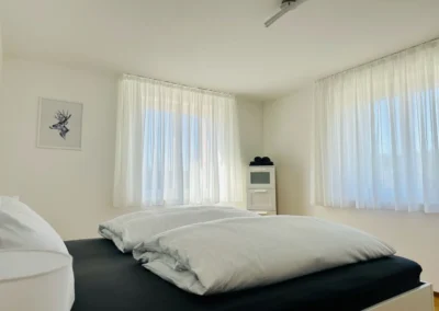 Schlafzimmer der Ferienwohnung Westweg mit großen Fenstern, hellen Vorhängen und Doppelbett.