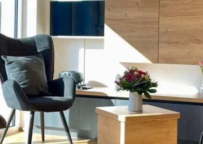 Wohnzimmerdetail der Ferienwohnung Westweg mit modernem Sessel, Blumengesteck und Holzsideboard.