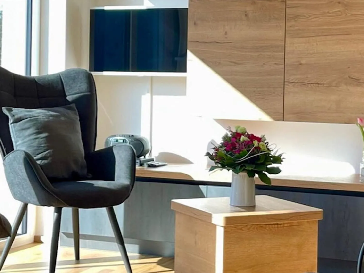 Wohnzimmerdetail der Ferienwohnung Westweg mit modernem Sessel, Blumengesteck und Holzsideboard.