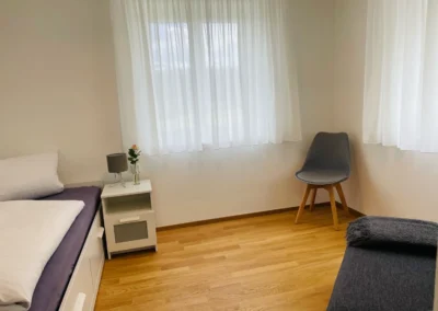 Zweites Schlafzimmer der Ferienwohnung Westweg mit Einzelbett, Nachttisch und grauem Stuhl.