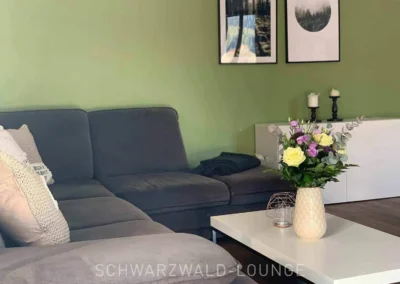 Gemütliches Wohnzimmer im Apartment Bergsee Schwarzwald mit grauem Sofa, Blumen auf dem Couchtisch und weißer Kommode vor grüner Wand.
