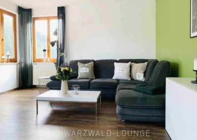 Wohnzimmeransicht im Apartment Bergsee Schwarzwald mit großer Sofalandschaft, Blumen und heller Fensterfront.