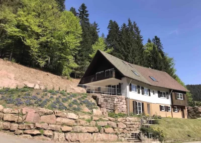 Hausansicht des Schwarzwald Chalet Lohmühle von der Straße mit Natursteinmauer und Waldkulisse.