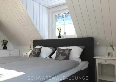 Helles Schlafzimmer im Schwarzwald Chalet Lohmühle mit Doppelbett, weißer Holzverkleidung und Blick durch das Fenster in die Natur.