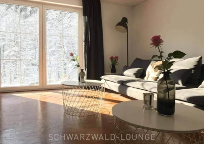 Geräumiges Wohnzimmer im Schwarzwald Chalet Lohmühle mit großem Panoramafenster, modernem grauen Sofa und Blick auf die verschneite Schwarzwaldlandschaft.