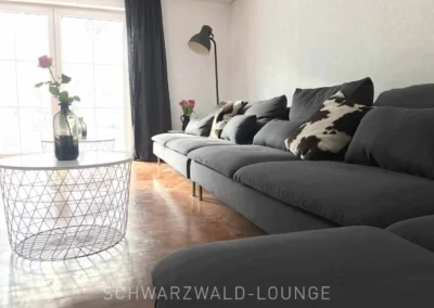 Modernes Wohnzimmer im Schwarzwald Chalet Lohmühle mit langem grauen Sofa, dekorativen Kissen und viel Tageslicht durch große Fenster.