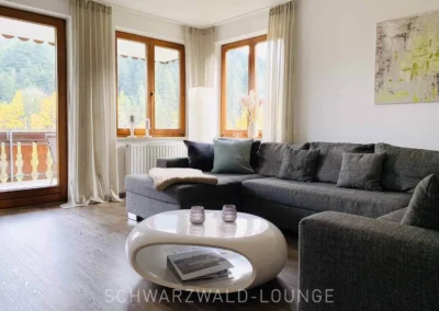 Wohnzimmer der Ferienwohnung Waldwiese mit großem Sofa und Panoramafenstern zur Schwarzwaldlandschaft.
